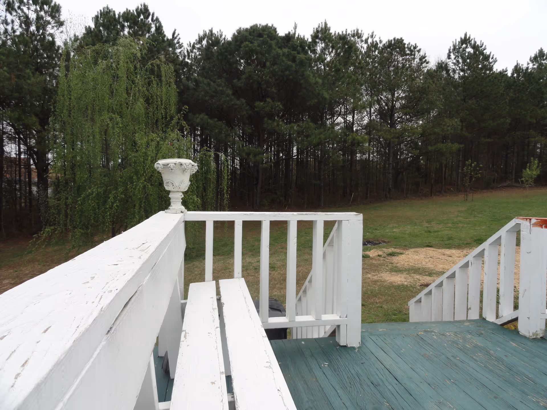 View from a wooden deck with white railings and a white planter on the corner, overlooking a grassy yard with trees in the background.
