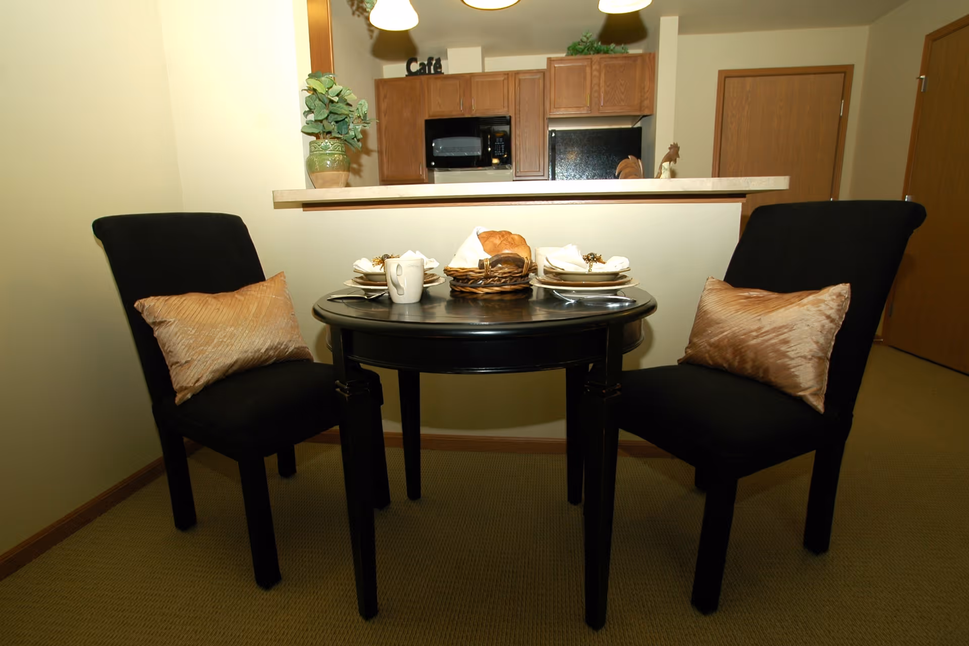 A small dining area with a round black table set for two, featuring plates, napkins, a mug, and a basket with bread. Two black chairs with beige cushions are positioned on either side of the table. In the background, there is a kitchen counter with wooden cabinets, a microwave, and a refrigerator. A potted plant and decorative items are on the counter and cabinets.