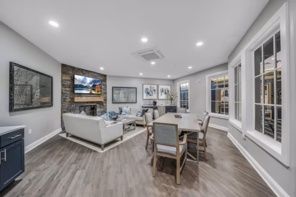 A modern living room with light gray walls and wood flooring. The room features a stone fireplace with a mounted flat-screen TV above it, two white sofas arranged around a coffee table, and a dining table with six chairs. Large windows with white frames allow natural light to enter the space. The room is decorated with framed artwork and has recessed ceiling lights.