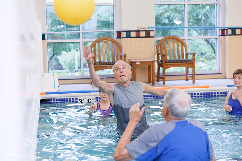 A group of elderly people in an indoor swimming pool playing with a large yellow ball. One man is reaching up to catch or hit the ball while others watch or participate. There are two wooden chairs and a small table by the window in the background.