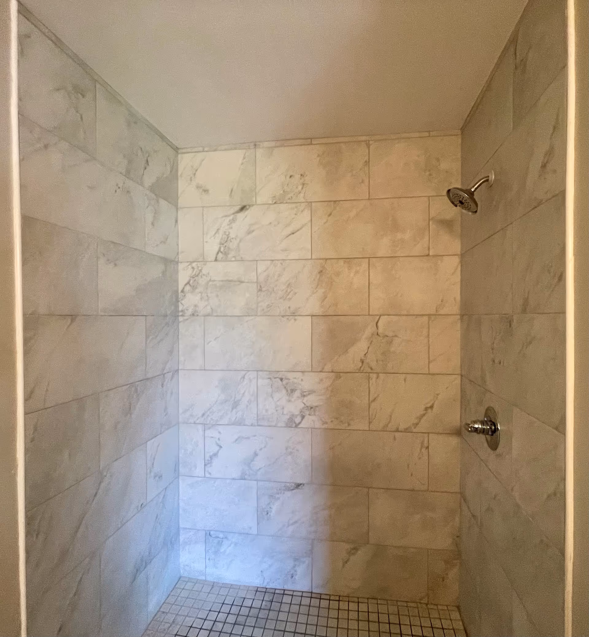 A tiled shower area with light-colored marble-patterned wall tiles and small square floor tiles. There is a silver showerhead and a control knob on the right wall.