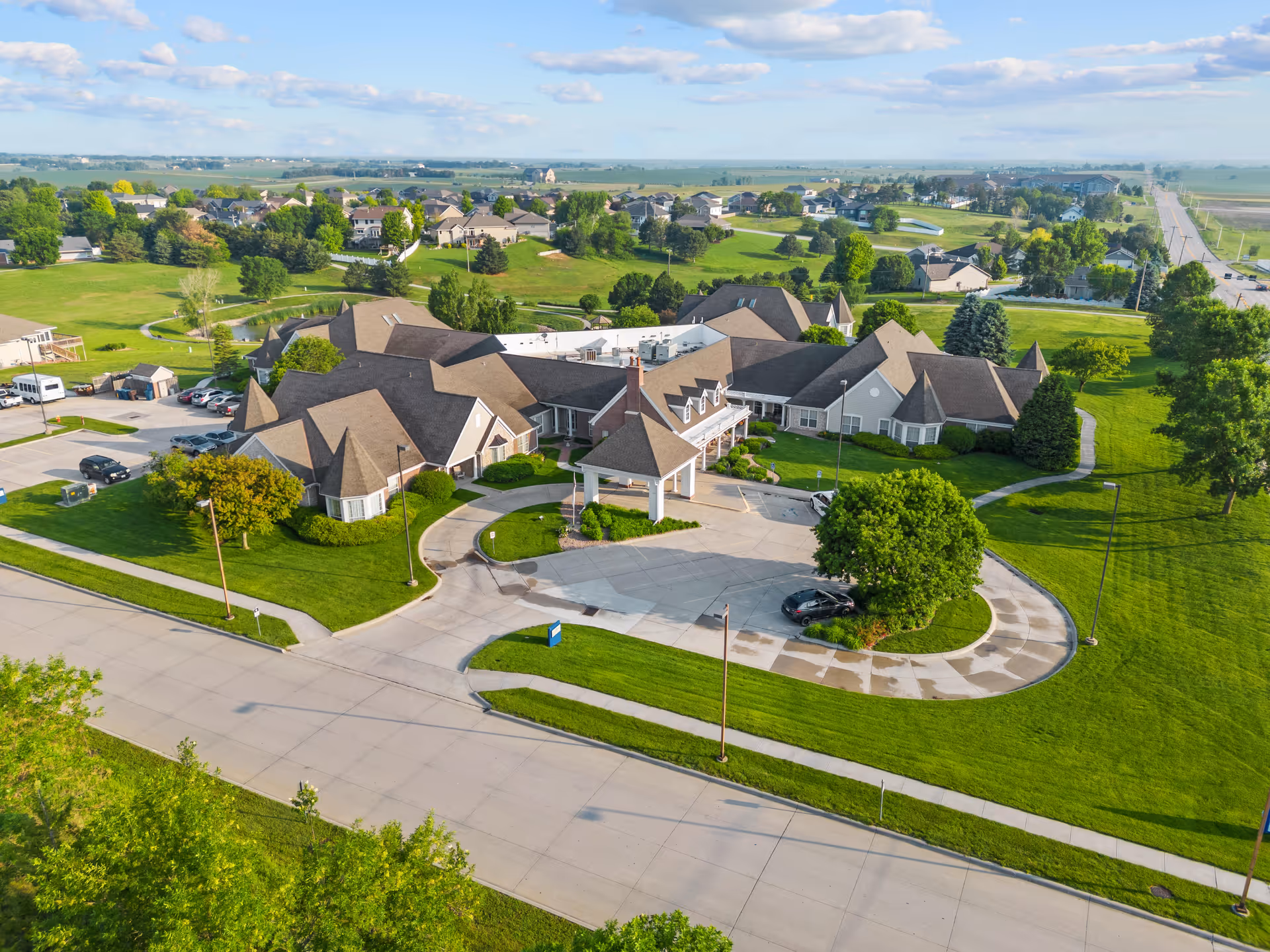 Aerial view of Good Samaritan Society - Prairie View Gardens facility surrounded by green lawns and trees, with a circular driveway and several parked cars. The building has multiple peaked roofs and is located in a suburban area with other houses and open fields in the background under a partly cloudy sky.