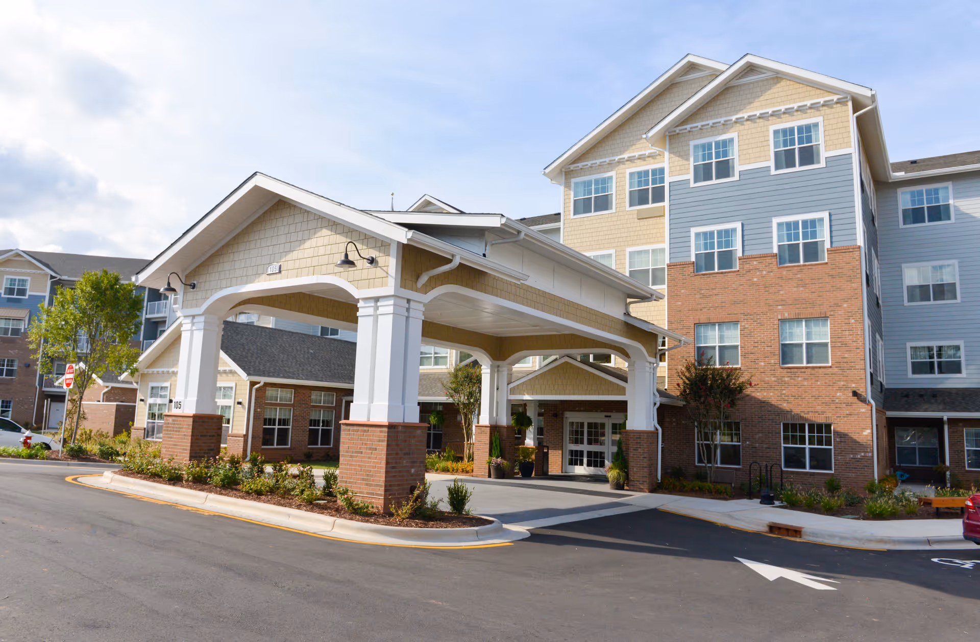 Exterior view of a multi-story senior living facility with a covered entrance supported by white columns and brick bases. The building features a combination of brick and siding in beige, blue, and white colors. There are landscaped areas with small bushes and trees around the entrance and parking lot.