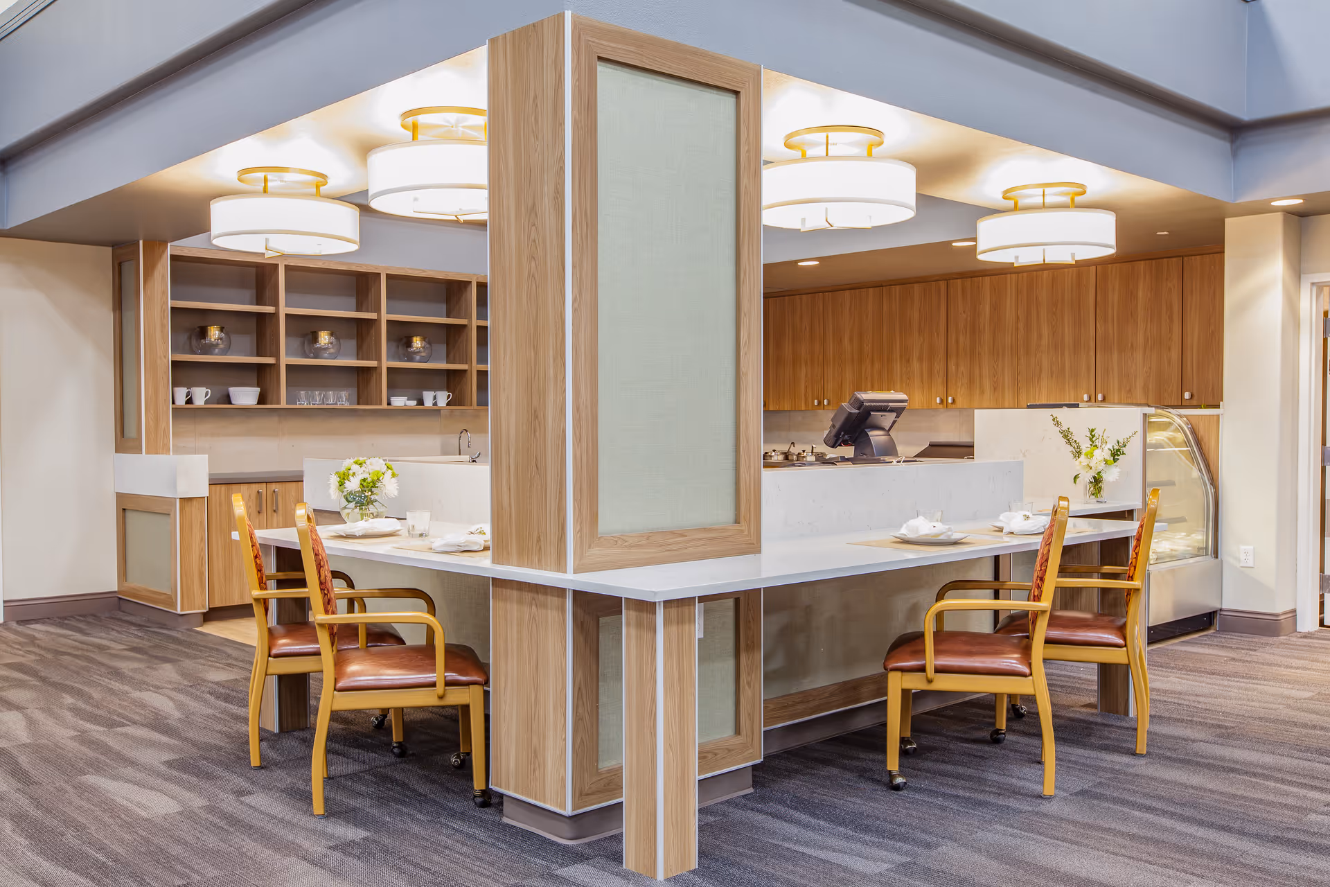 A modern dining area with a central wooden column surrounded by a white countertop. Four wooden chairs with brown leather seats are placed around the counter, each set with white plates and napkins. The background features wooden cabinets, open shelves with cups and glasses, and a small display refrigerator. The ceiling has multiple round, white light fixtures.