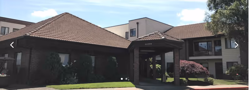Exterior view of King City Senior Village showing a building with a brown tiled roof, brick and light-colored siding, surrounded by greenery and trees under a blue sky with some clouds.
