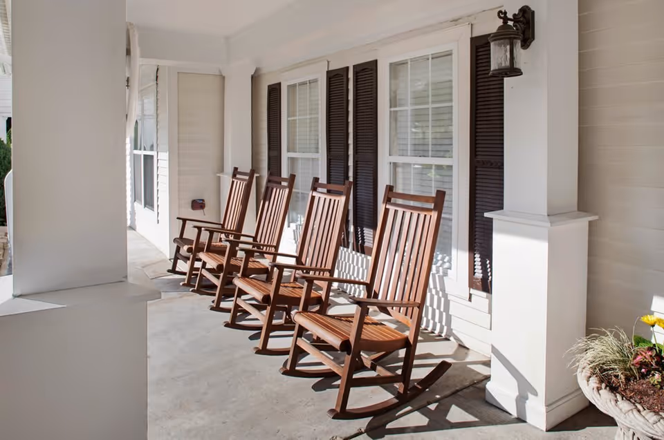 A row of five wooden rocking chairs lined up on a covered porch with white pillars and windows with dark shutters. There is a wall-mounted lantern light fixture and a planter with flowers on the right side.
