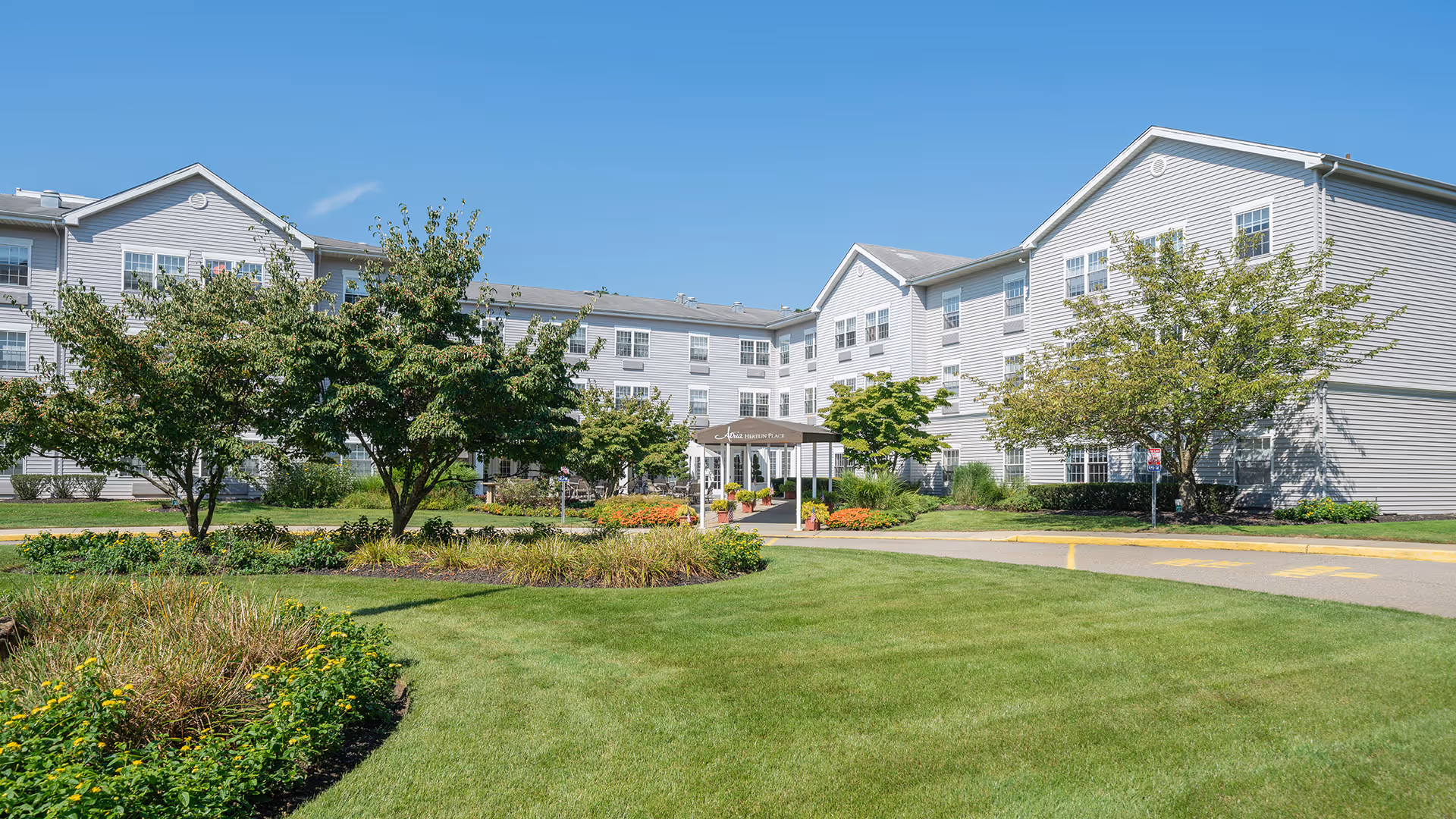 Exterior view of Atria Hertlin Place, a three-story senior living facility with light gray siding, multiple windows, and a covered entrance. The building is surrounded by well-maintained green lawns, trees, and flower beds under a clear blue sky.