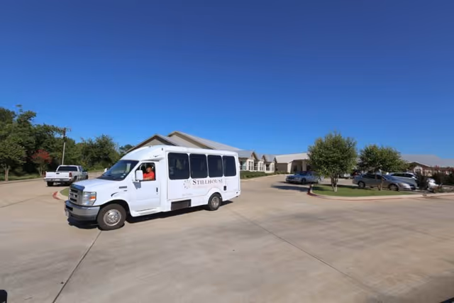 A white shuttle van with the logo 'Stillhouse' on the side is parked on a wide concrete driveway in front of a single-story building with a metal roof. Several cars are parked near the building, and there are small trees and greenery around the parking area under a clear blue sky.