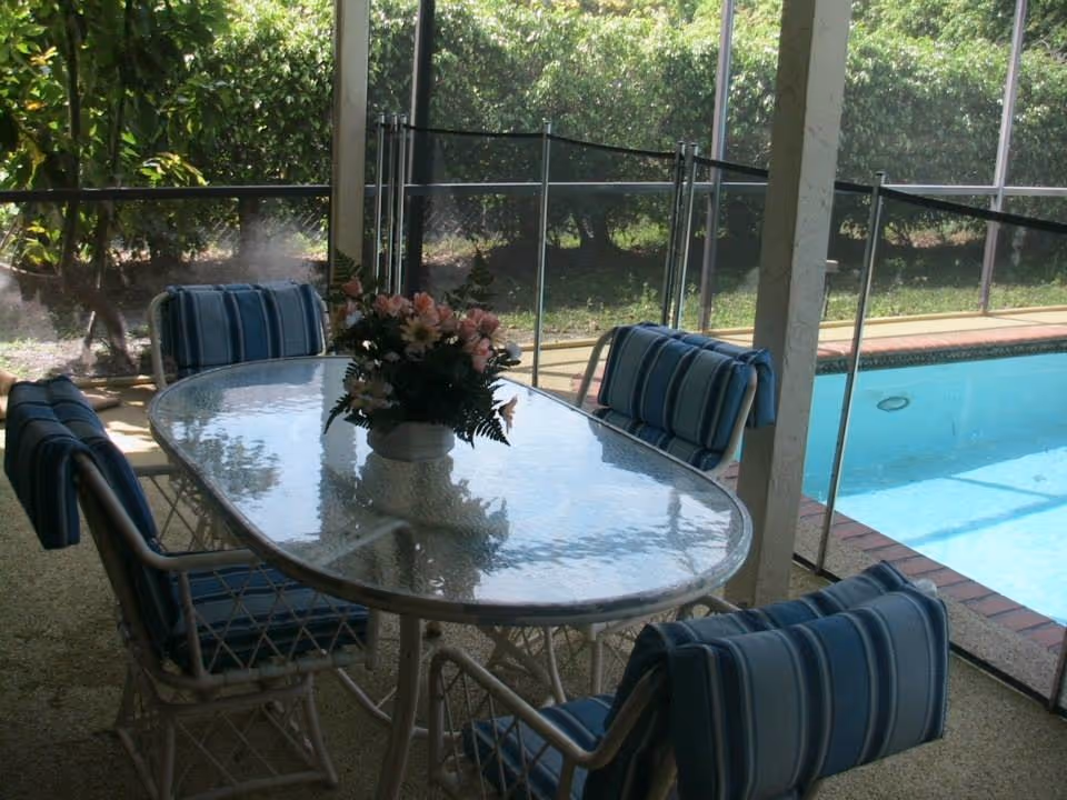 Outdoor patio with glass table, blue cushioned chairs, and a pool behind a screened enclosure.