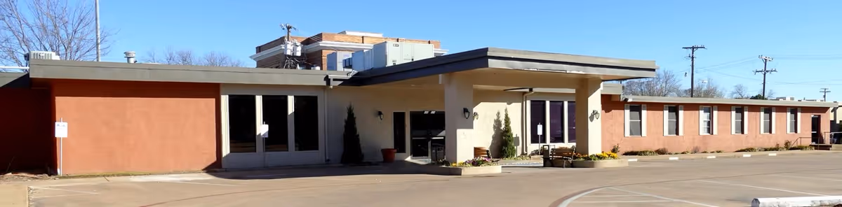 Exterior view of a single-story healthcare facility building with a flat roof, beige and reddish-brown walls, several windows, and a main entrance with glass doors. The building is surrounded by a paved parking area with clear skies above.