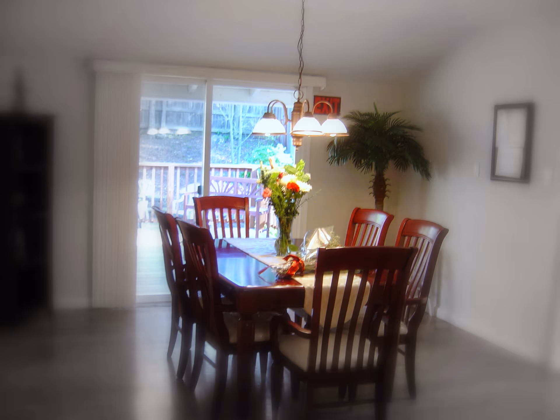 Dining room with a wooden table and chairs, a hanging light fixture, a vase of flowers, sliding glass doors to a deck, and a potted plant.