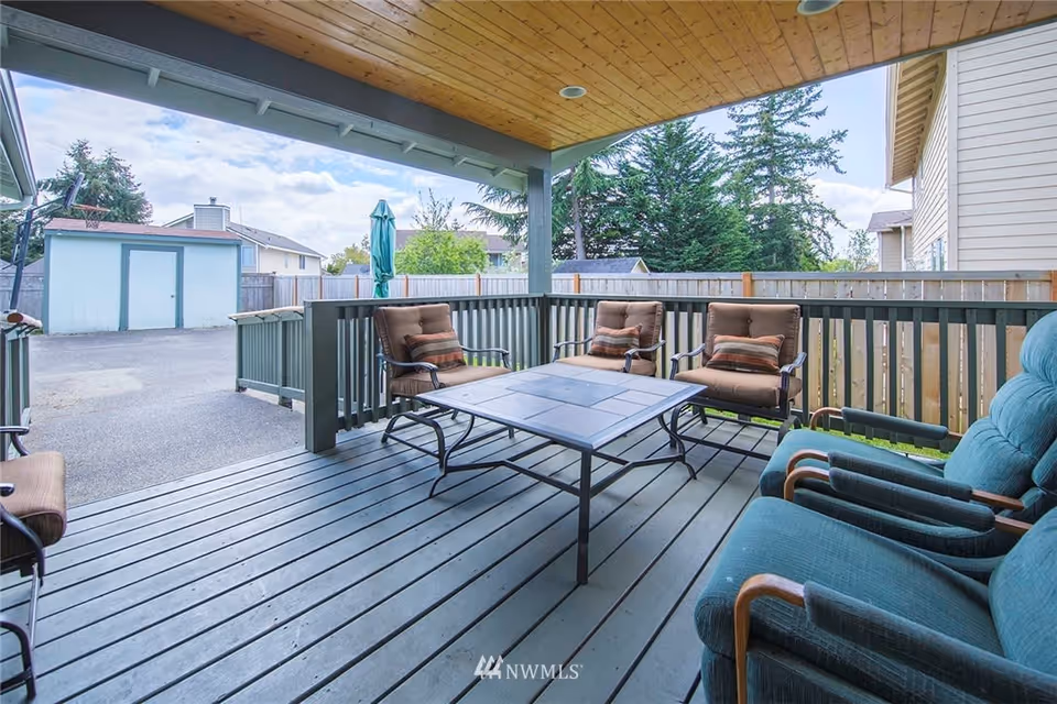Covered backyard deck with outdoor seating and a central table overlooking a fenced yard and storage shed.