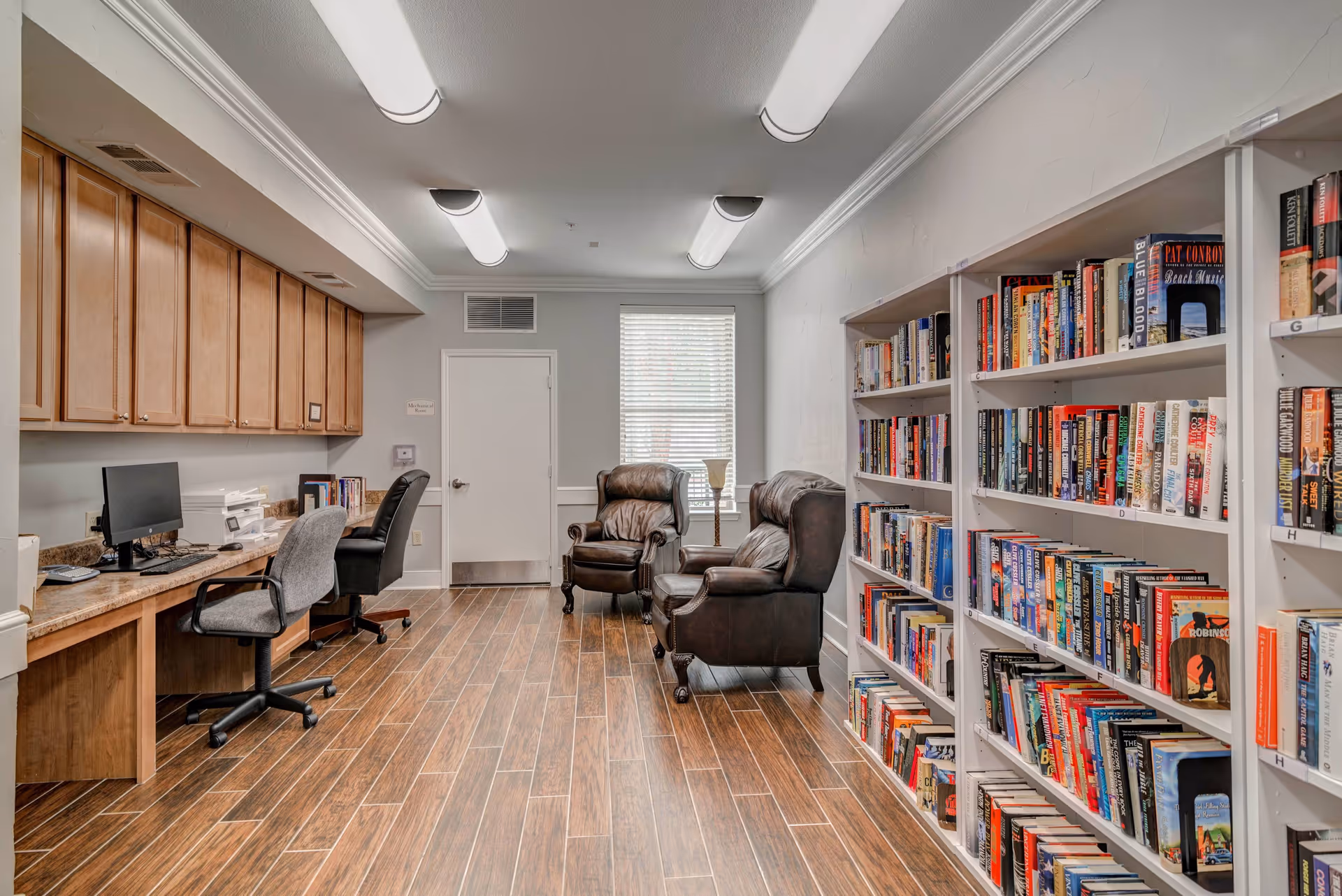 A bright community reading room with bookshelves along the right wall, two leather armchairs by a window, and a desk area with computers on the left.