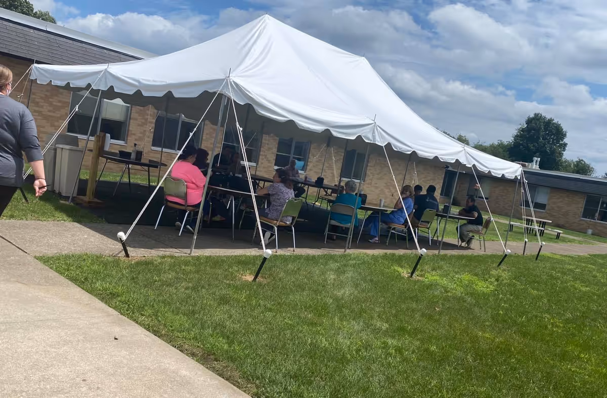 Several people sitting under a large white canopy tent outside a brick building on a sunny day. The tent is set up on a concrete area surrounded by green grass, and the people appear to be engaged in conversation or an activity.
