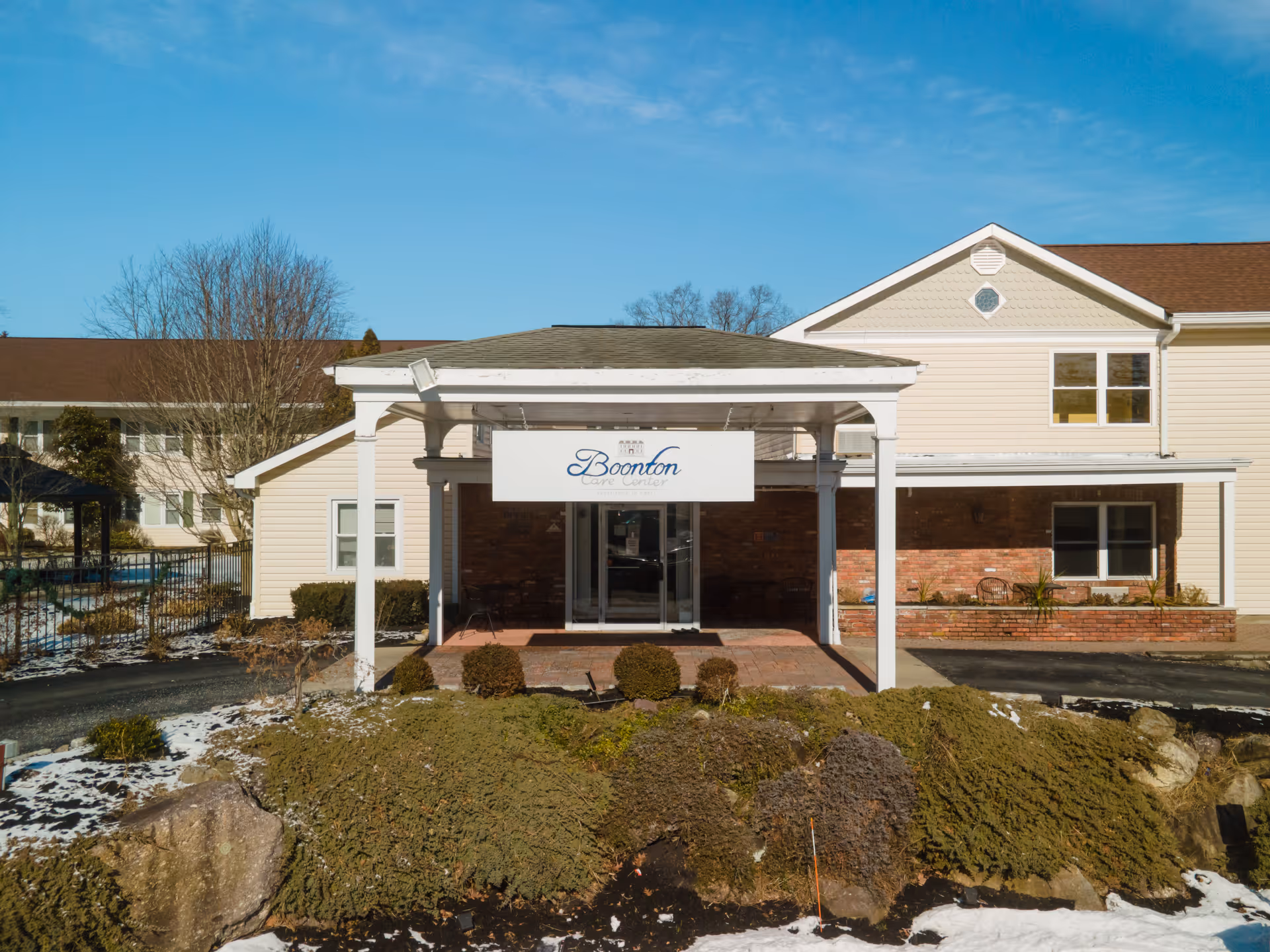 Covered entrance and porte-cochère of a beige two-story nursing home building with a sign above the glass doors and landscaped grounds with light snow.