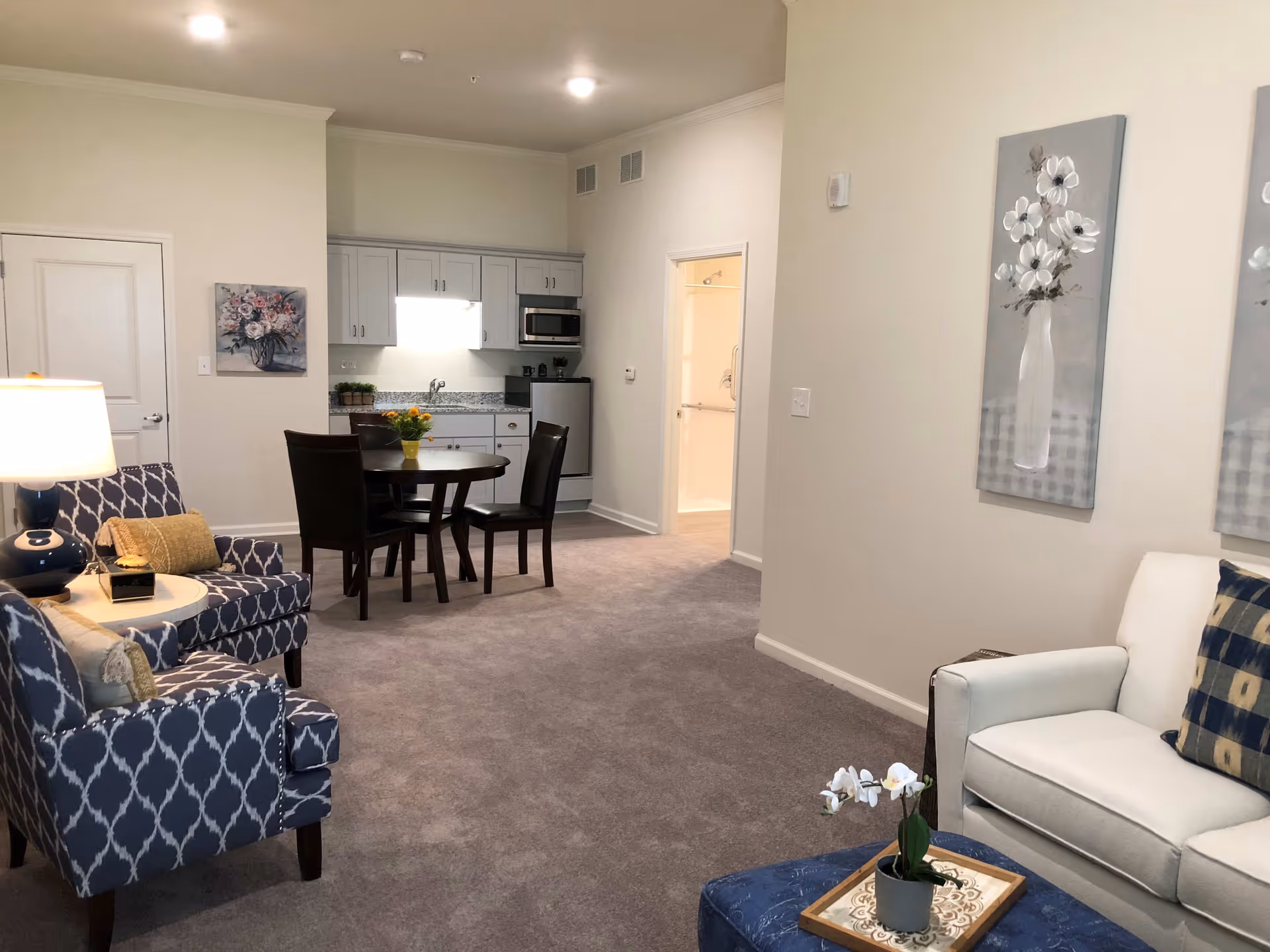 A cozy living area with a white sofa and a blue patterned armchair, a small round dining table with four chairs, and a compact kitchen with white cabinets, a microwave, and a mini fridge. The room has beige walls, carpeted floor, and floral artwork on the walls. A doorway leads to a bathroom with a visible shower.