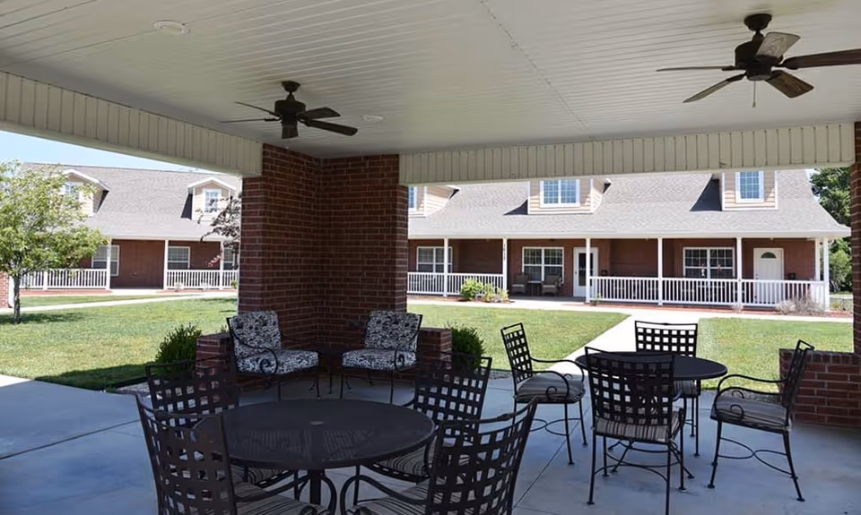 Covered outdoor seating area with metal tables and chairs, ceiling fans, and a view of a grassy courtyard and brick senior living buildings with white railings and dormer windows.