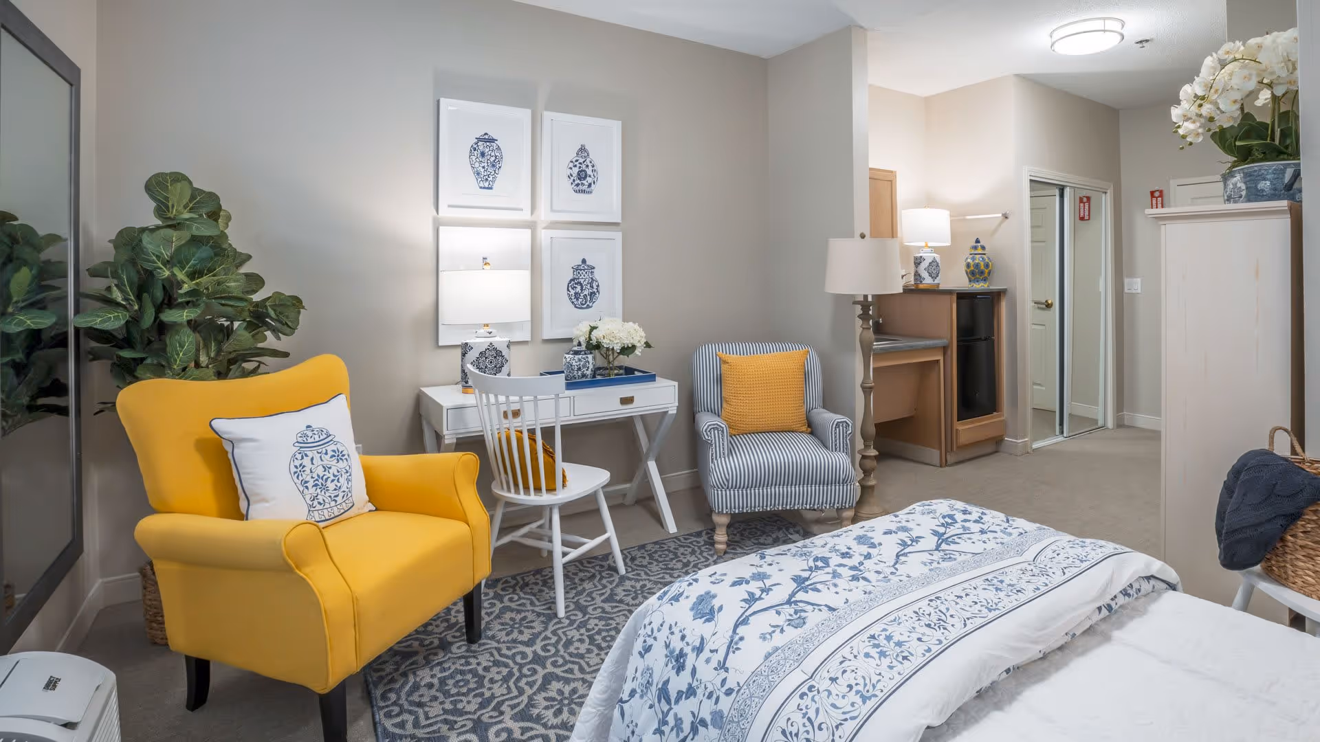 A cozy bedroom featuring a bed with blue and white floral bedding, a yellow armchair with a decorative pillow, a striped armchair with a yellow cushion, a white desk with a chair, and framed blue and white artwork on the wall. The room also includes a tall floor lamp, a large mirror, a potted plant, and a cabinet with a vase of white flowers.