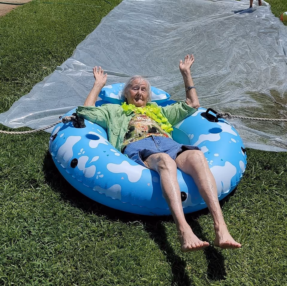 An elderly woman wearing a green shirt, floral top, blue shorts, and a yellow lei is sitting on a blue and white inflatable tube on a grassy lawn. She has her arms raised and appears to be enjoying a slip-and-slide activity with a large plastic sheet behind her.