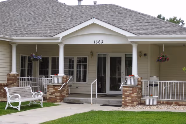 Front exterior view of a single-story building with a covered porch supported by white columns. The porch has brick pillars with white railings and hanging flower baskets. There is a white bench on the lawn near the sidewalk leading to the entrance, which has double glass doors and the number 1663 above them.