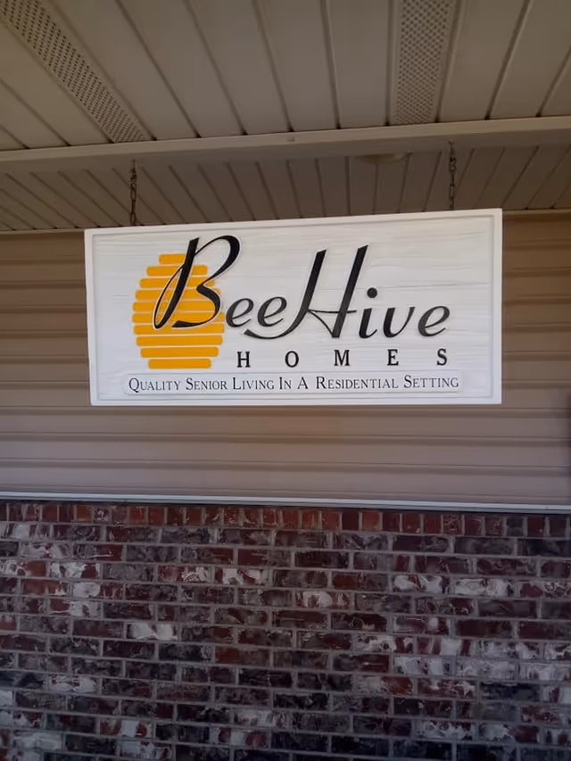 A hanging sign for Bee Hive Homes with a yellow beehive graphic and the text 'Quality Senior Living In A Residential Setting' beneath the name. The sign is mounted under a roof with beige siding and a brick wall below.