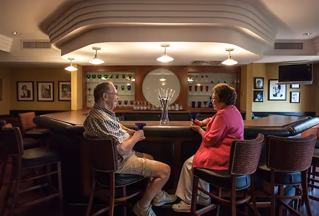 An elderly man and woman sit on bar stools facing each other at a bar counter in a cozy room. They are holding blue glasses and appear to be engaged in conversation. Behind the bar, there are shelves displaying various glassware and a large decorative glass centerpiece. The room is warmly lit with ceiling lights and decorated with framed black and white photos on the walls.