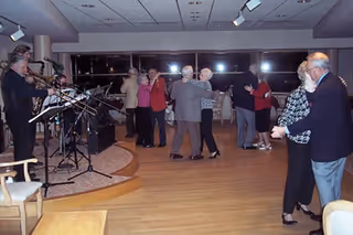 A group of elderly people dancing in pairs on a wooden floor in a well-lit room with large windows. A live band is playing music on a small stage to the left side of the image. The atmosphere appears lively and social.
