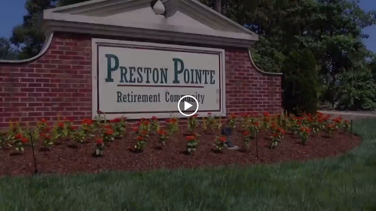 A brick sign for Preston Pointe Retirement Community surrounded by a flower bed with red flowers and green grass, with trees and a clear blue sky in the background.