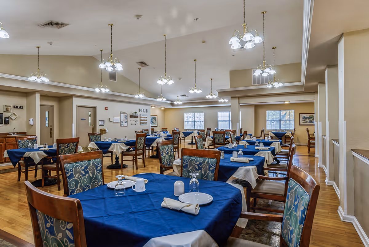 A spacious dining room with multiple tables covered in blue tablecloths and white napkins. Each table is set with plates, glasses, and utensils. The room has wooden floors, beige walls, and several hanging light fixtures. Windows along the far wall let in natural light.