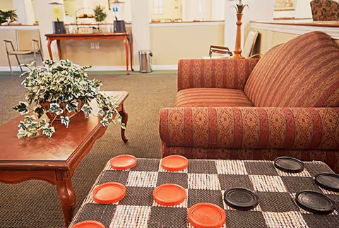 A cozy living room area with a patterned red armchair, a wooden coffee table with a green leafy plant, and a checkers board with red and black pieces set up on a table. In the background, there is a wooden console table with decorative items and a chair against a light-colored wall.