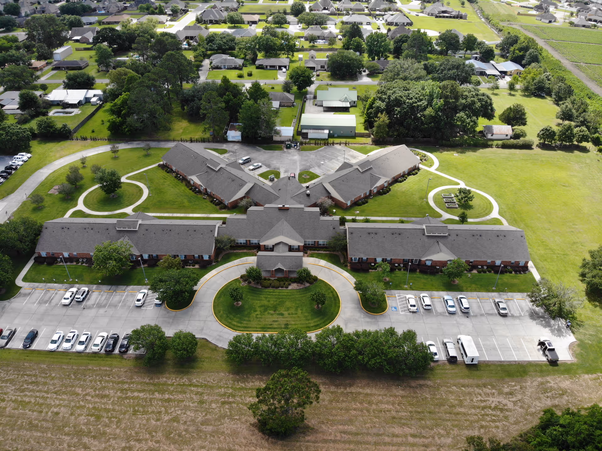 Aerial view of Azalea Estates of New Iberia, showing a large, single-story senior living facility with multiple wings arranged in a symmetrical layout. The building is surrounded by well-maintained green lawns, circular walking paths, and parking lots with several cars parked. Residential houses and trees are visible in the background.