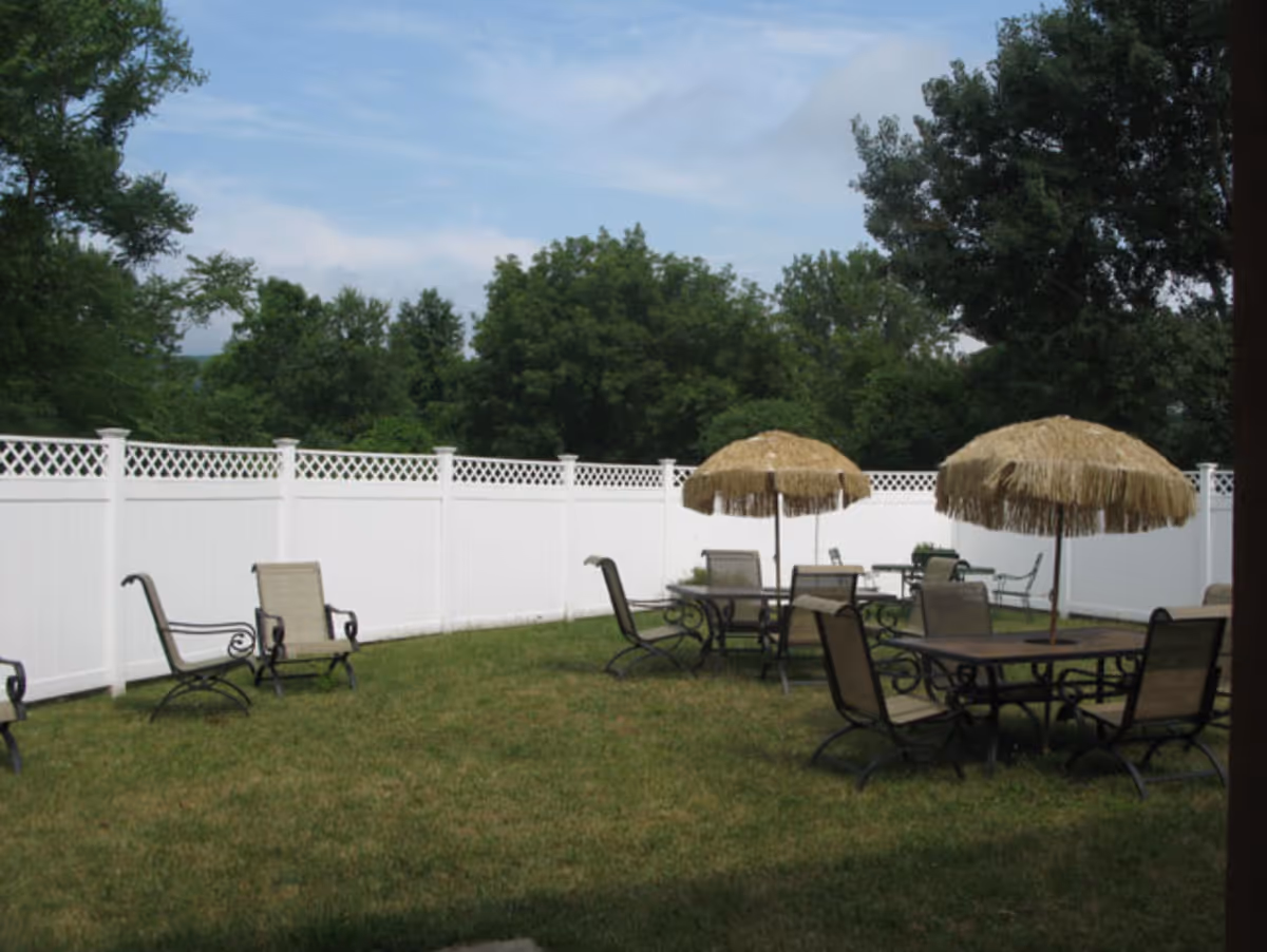 Outdoor seating area with multiple tables and chairs on a grassy lawn, each table shaded by thatched umbrellas, surrounded by a white privacy fence and trees in the background under a partly cloudy sky.