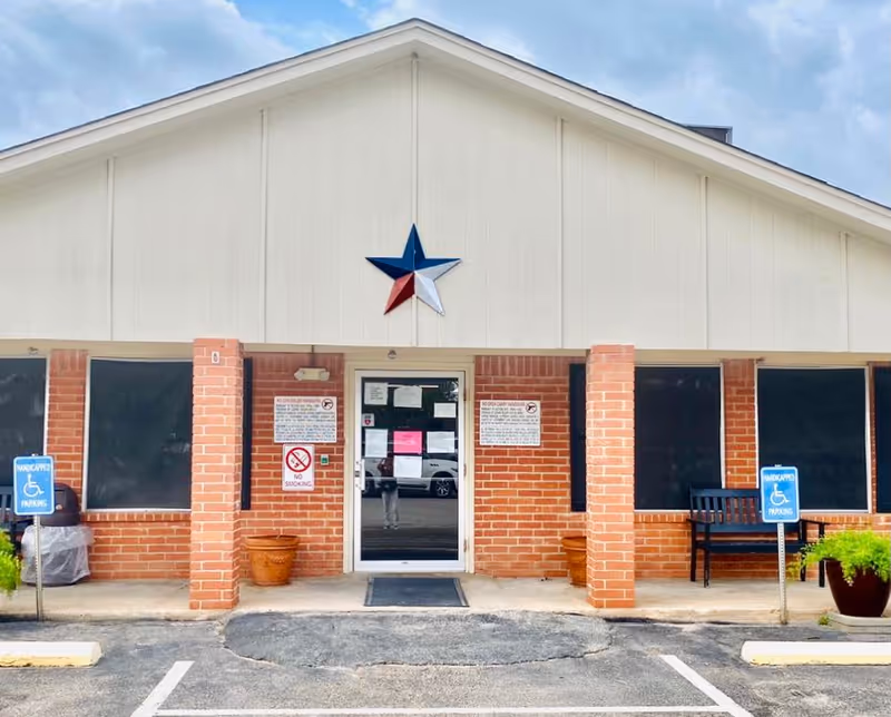 Front exterior view of Concho Health and Rehabilitation Center showing a brick building with a white upper facade and a large Texas star decoration above the entrance door. There are two handicapped parking signs and a bench near the entrance.