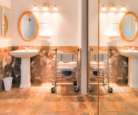 Accessible bathroom with pedestal sinks, oval mirrors, and a wheeled shower/commode chair reflected in a large mirror.