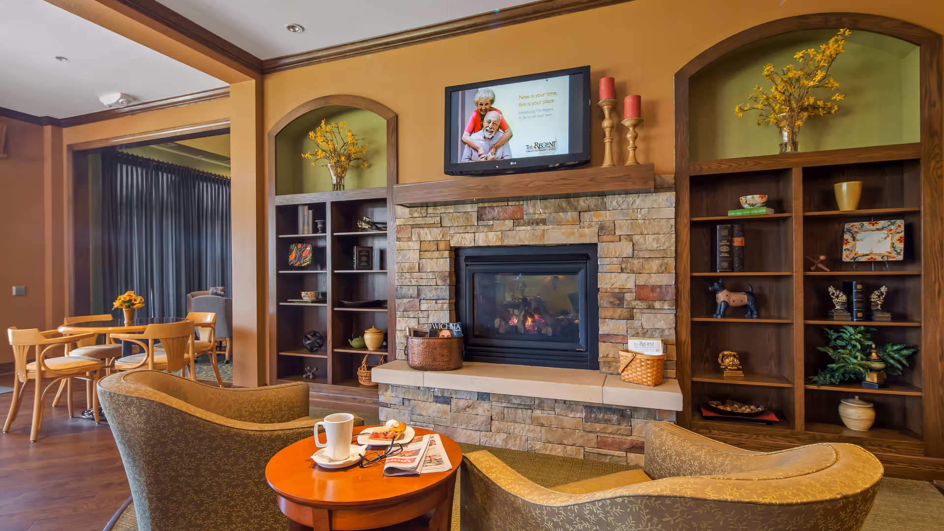 Cozy senior living lounge with a stone fireplace flanked by built-in shelves, two armchairs and a small table holding a cup and magazines.