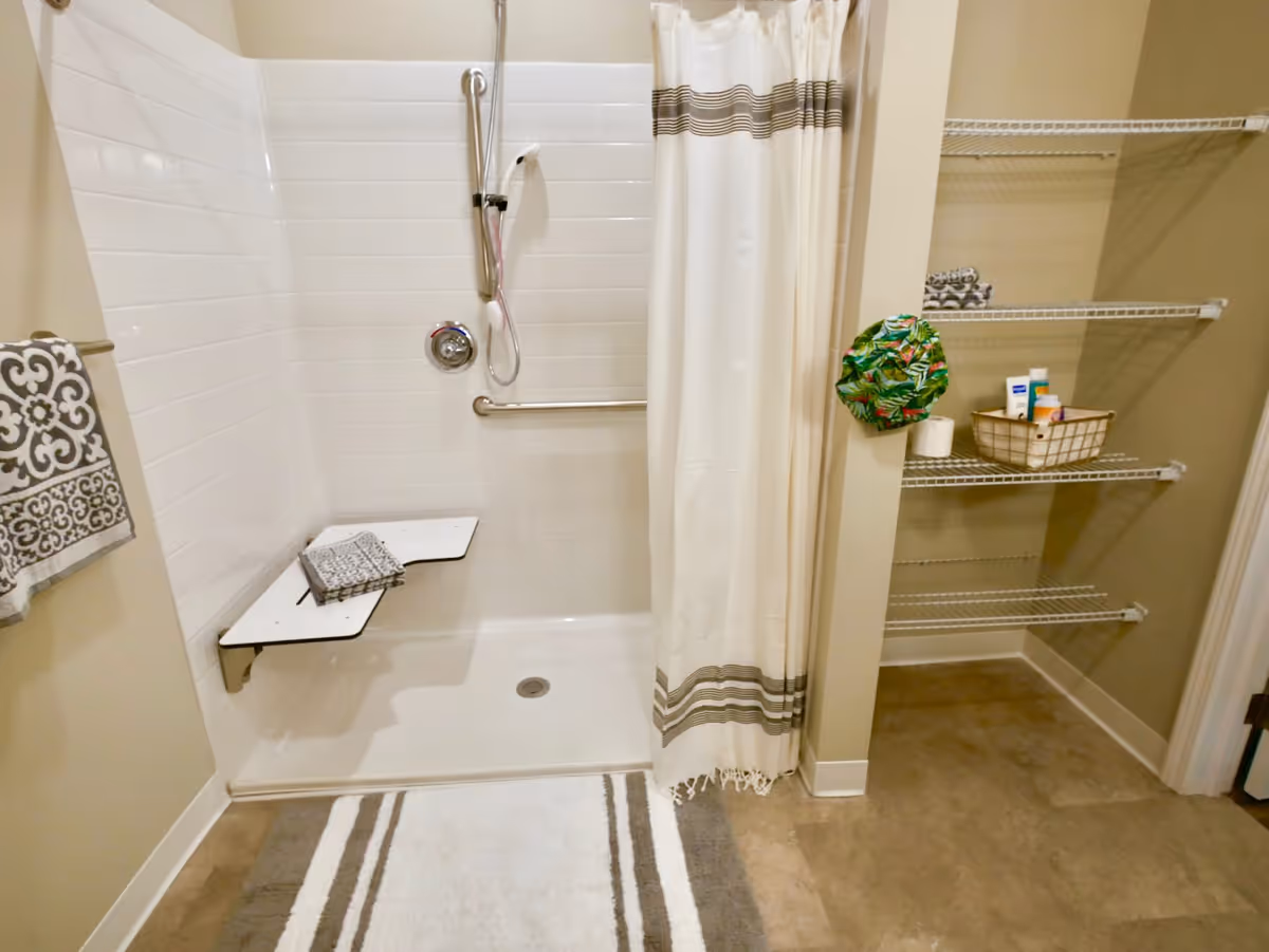 A bathroom shower area with white tiled walls and a white shower curtain with brown stripes. There is a foldable shower seat with a folded towel on it, a handheld showerhead, and grab bars for support. To the right, there are wire shelves holding toiletries and a green patterned shower cap hanging on the wall. A patterned towel hangs on a towel rack to the left, and a striped bath mat is on the floor in front of the shower.