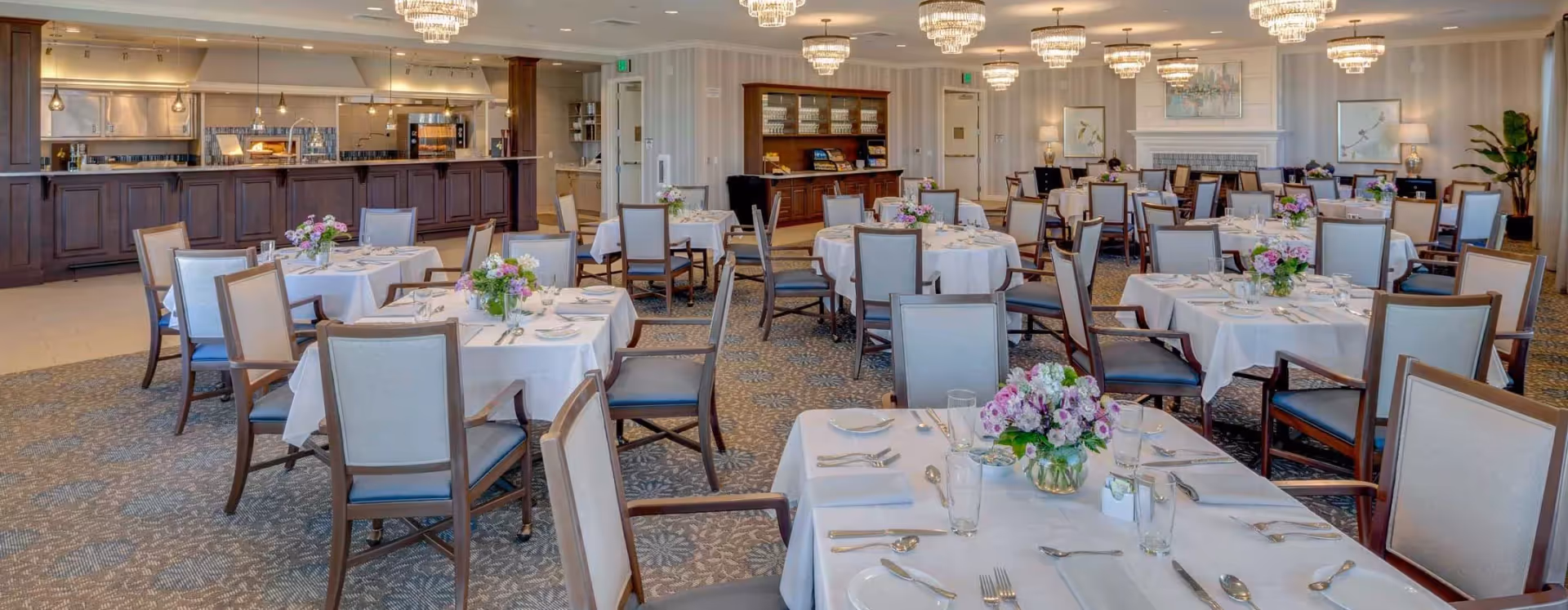 A spacious and elegant dining room with multiple round tables covered with white tablecloths, each set with plates, glasses, and silverware. The tables are decorated with floral centerpieces. The room features a carpeted floor, chandeliers hanging from the ceiling, a fireplace, and a serving counter with kitchen appliances in the background.