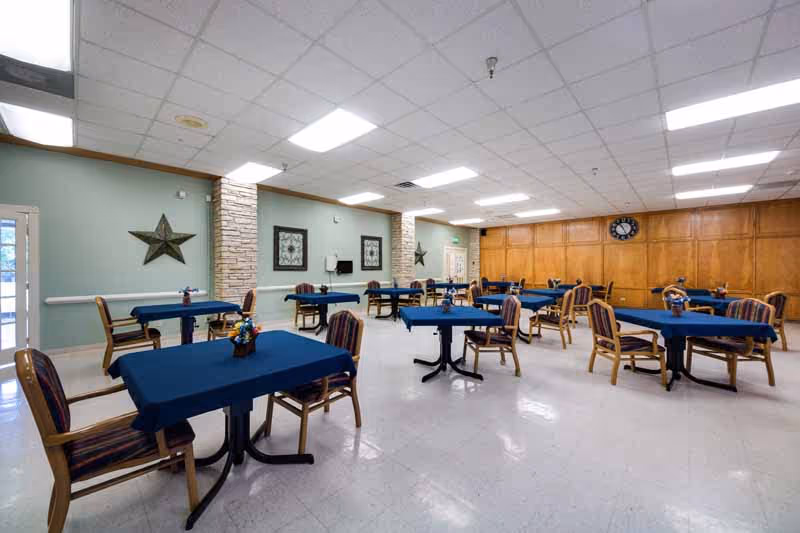 A spacious dining room with multiple tables covered in blue tablecloths, each surrounded by wooden chairs with striped cushions. The room has a light green wall with decorative stars and framed artwork, a clock on a wooden paneled wall, and bright overhead lighting.