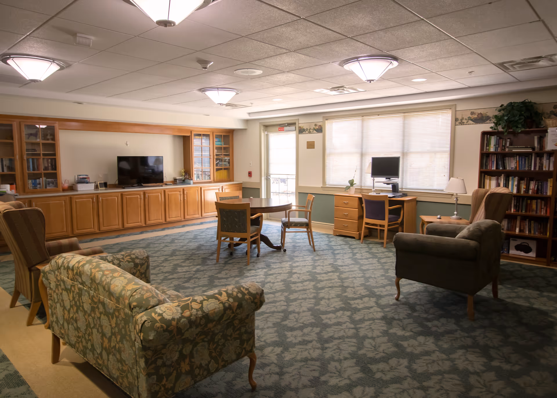 A cozy living room area in a senior living facility with patterned armchairs and a sofa, a round table with chairs in the center, a TV on a wooden cabinet along the wall, a desk with a computer near a window, and bookshelves filled with books. The room has soft lighting from ceiling fixtures and a carpet with a leaf pattern.