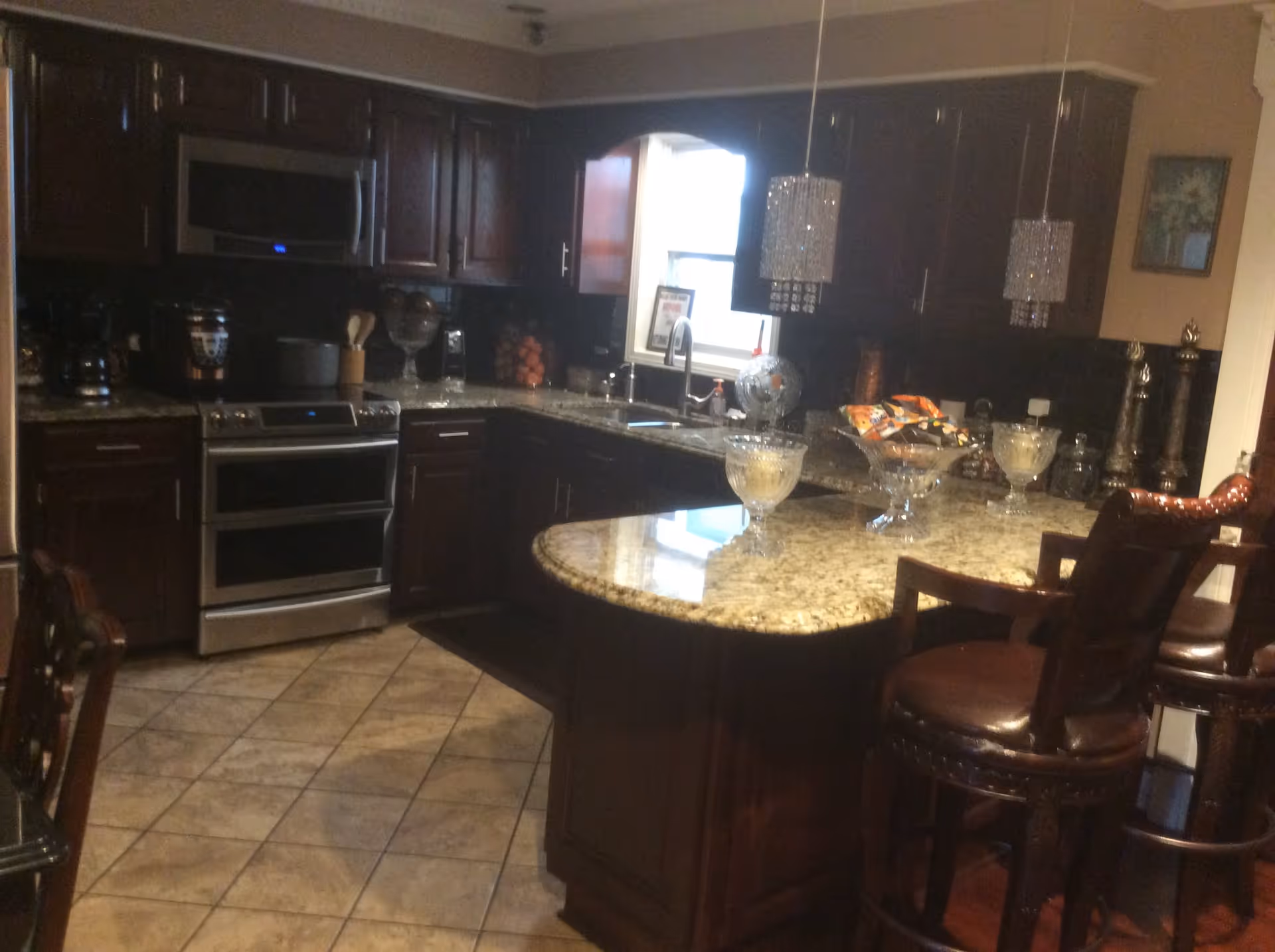 A kitchen with dark wooden cabinets, granite countertops, and tiled floor. There is a stainless steel oven and microwave, a sink under a window, and three crystal pendant lights hanging above a curved kitchen island with three brown leather bar stools. Various kitchen items and decorative bowls are placed on the countertops.