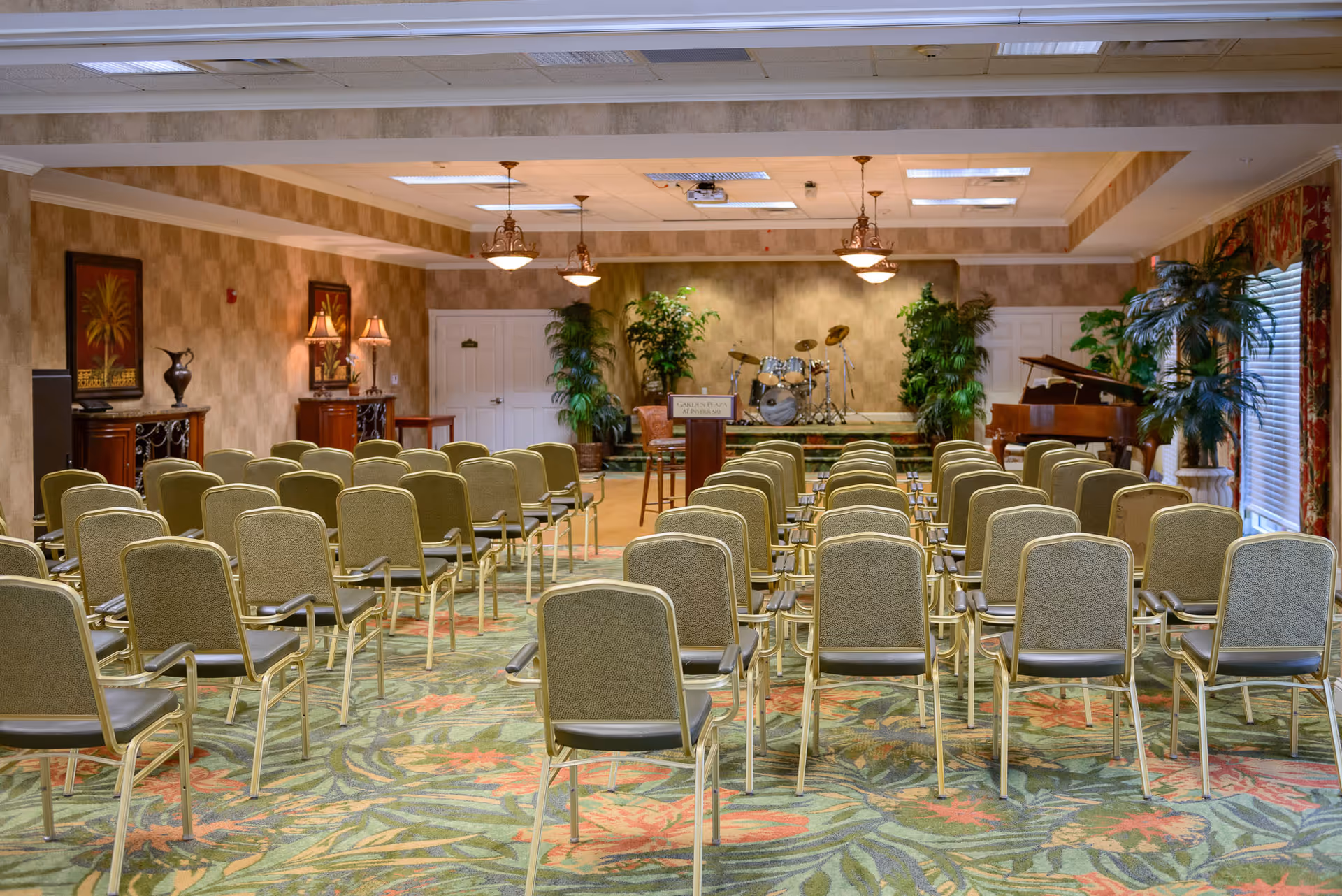 A conference or event room with rows of green cushioned chairs facing a small stage. The stage has a drum set, a podium with a sign that reads 'Garden Plaza at Inverrary,' and is flanked by large potted plants. The room has patterned carpet, warm lighting fixtures hanging from the ceiling, framed artwork on the walls, and a grand piano near a window with blinds and floral curtains.