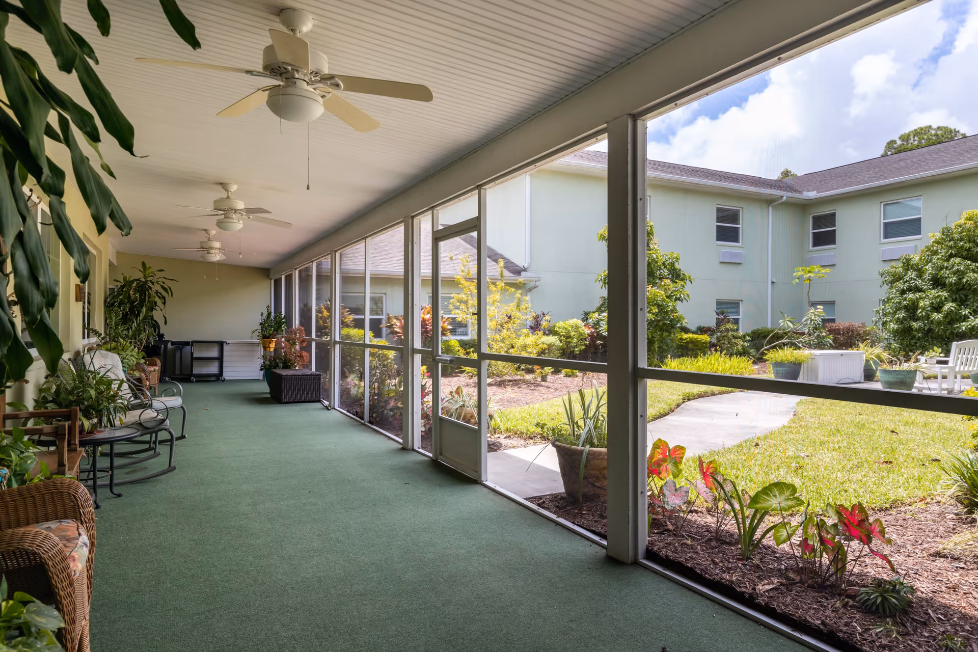 A screened-in porch area with green carpet flooring, ceiling fans, and various potted plants and wicker chairs. The porch overlooks a garden with grass, shrubs, and a light green building in the background under a partly cloudy sky.
