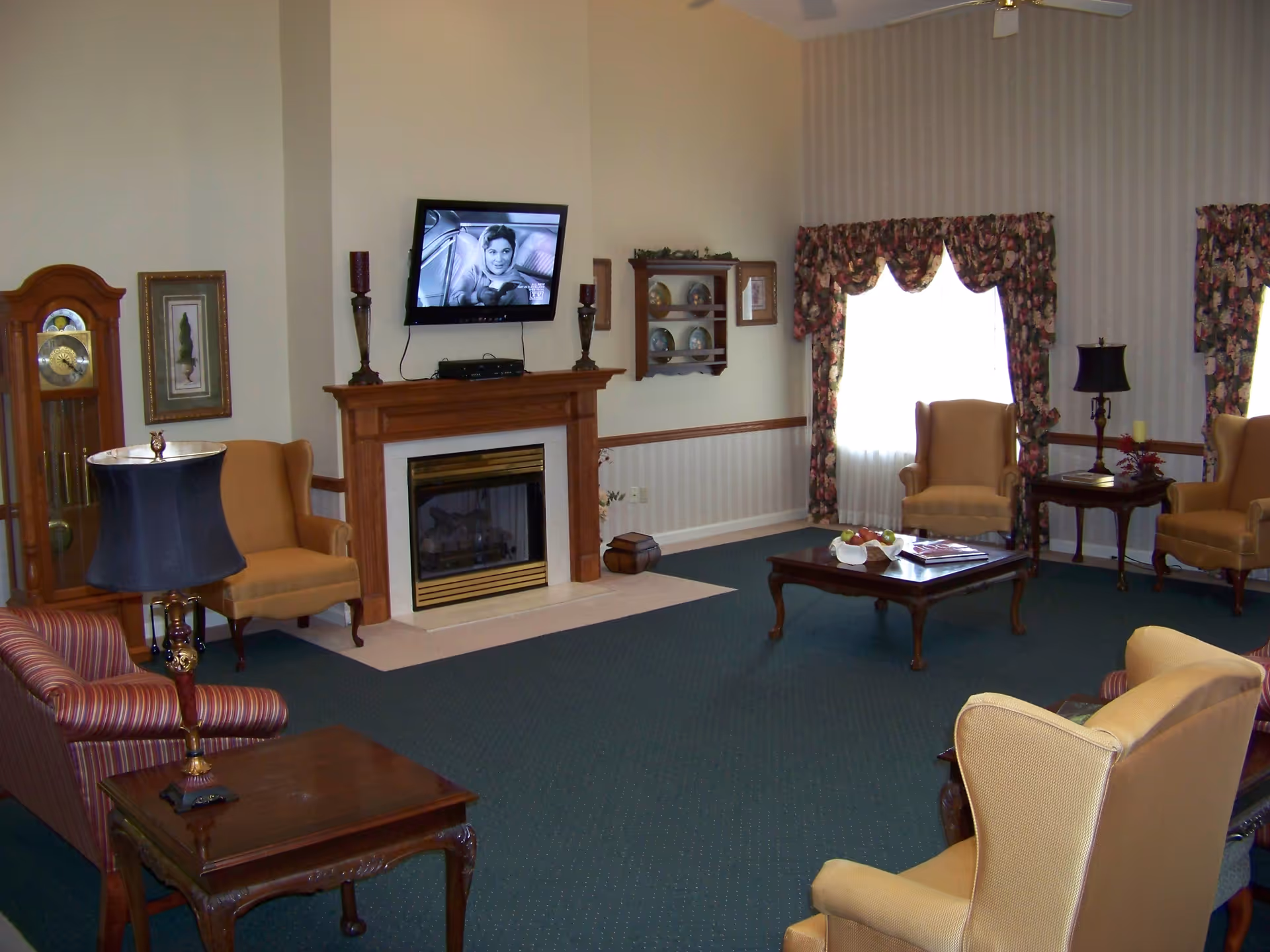 Comfortable memory care living room with a fireplace, wall-mounted TV, armchairs, coffee table and grandfather clock.