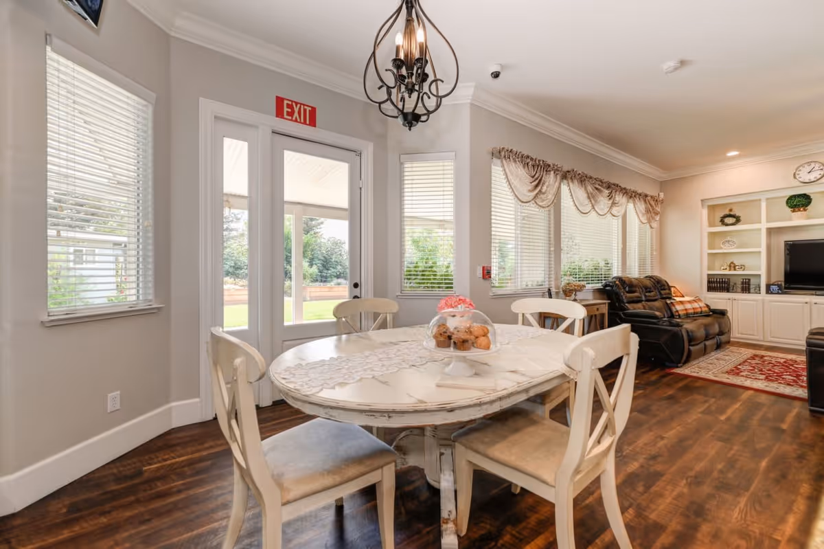 A cozy living and dining area with a round white wooden table surrounded by four chairs. On the table, there is a glass dome covering muffins. The room has large windows with blinds and decorative valances, a dark leather reclining sofa, a red patterned rug, built-in white shelves with books and decor, and a TV. A chandelier hangs above the dining table, and a glass door with an exit sign leads outside to a green lawn.