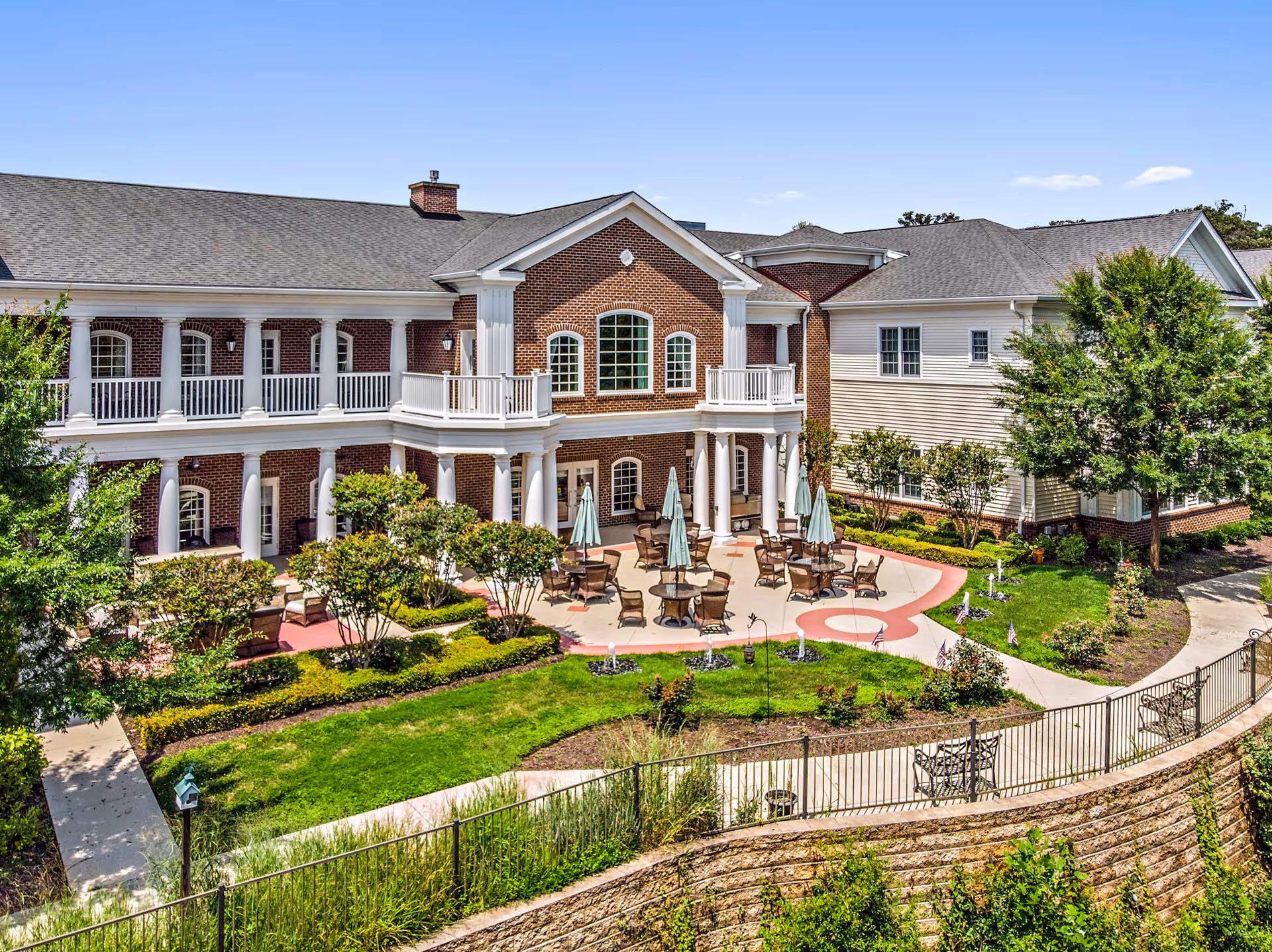 Outdoor patio area of a senior living facility with multiple tables and chairs under closed umbrellas, surrounded by well-maintained gardens and greenery. The building features red brick and white siding with large windows and white columns supporting a covered balcony.