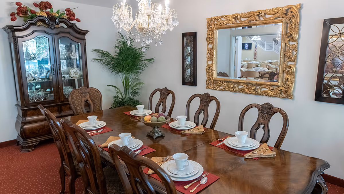 A formal dining room with a long wooden table set for six with white plates, cups, and silverware on red and beige placemats. A decorative bowl with artificial fruit is centered on the table. The room features a large ornate gold-framed mirror on the wall, a crystal chandelier hanging above the table, a tall wooden china cabinet filled with glassware and dishes, and a green potted plant in the corner.