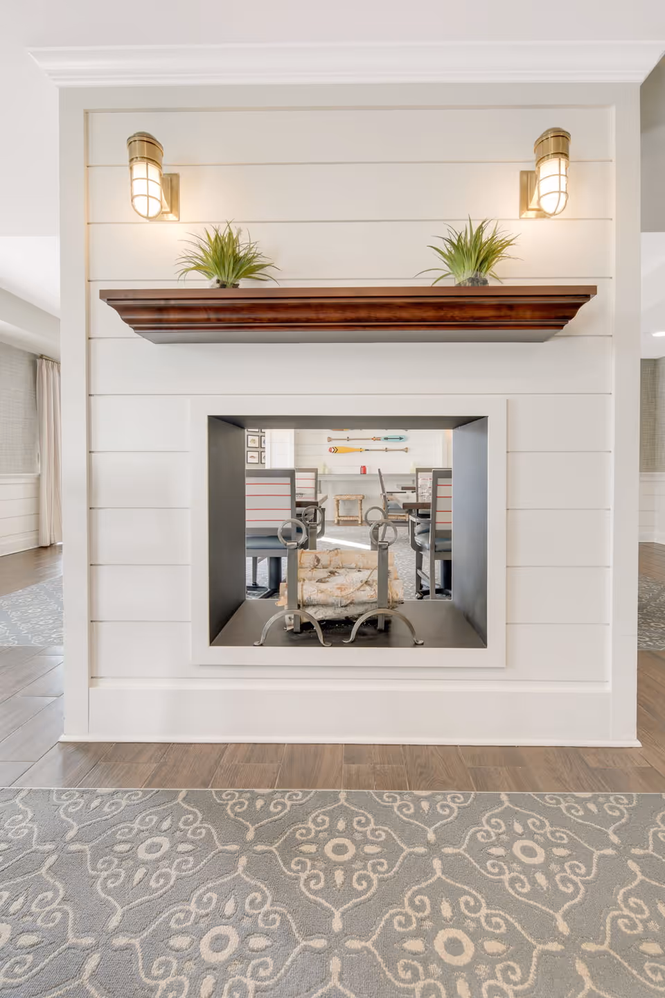A modern double-sided fireplace with a white shiplap surround and a wooden mantel holding two small potted plants. The fireplace is situated in a room with patterned carpet and wooden flooring, with a dining area visible through the fireplace opening.