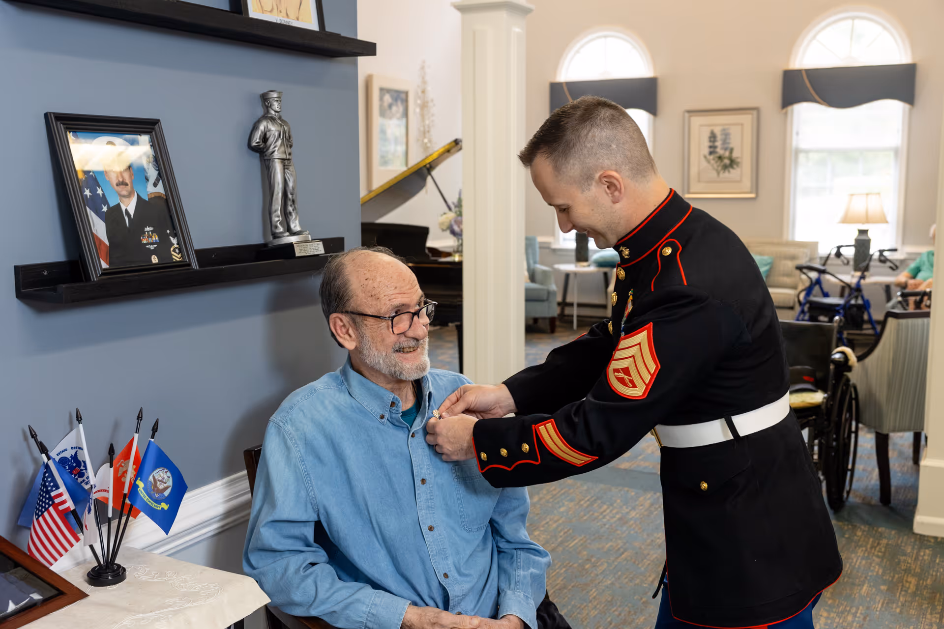 A young man in a military dress uniform pins a medal on an elderly man wearing glasses and a blue shirt, who is seated and smiling. The setting appears to be a senior living facility with a piano, framed photos, small flags, and comfortable seating in the background.