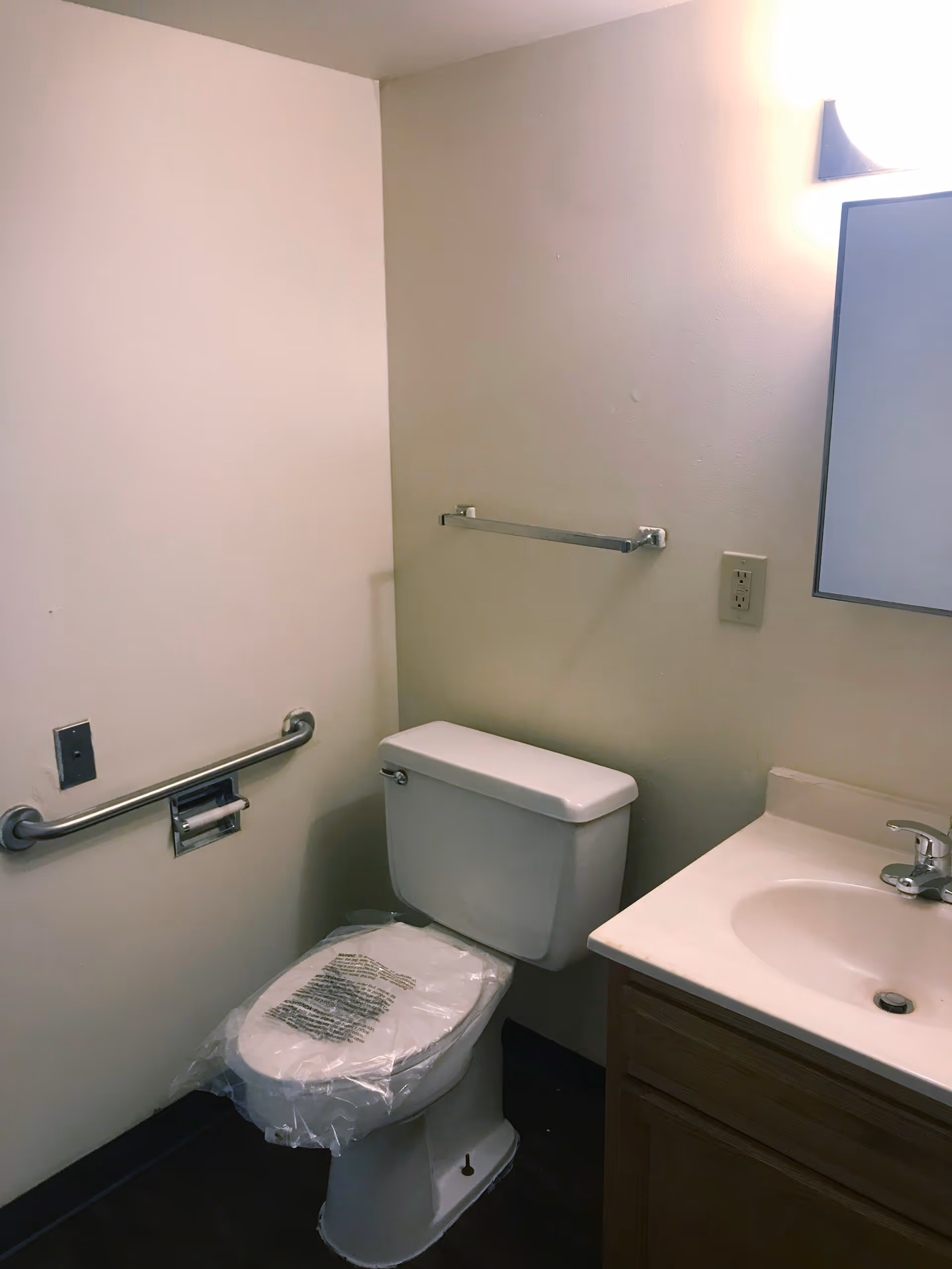 A small bathroom with a toilet covered in protective plastic, a sink with a faucet, a mirror above the sink, a towel rack on the wall, and a grab bar next to the toilet.