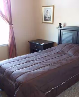 A small bedroom with a dark bed, nightstand, purple curtains, and framed art.